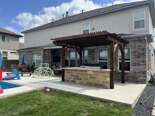 Stained cedar pergola over a stone outdoor kitchen beside a backyard pool